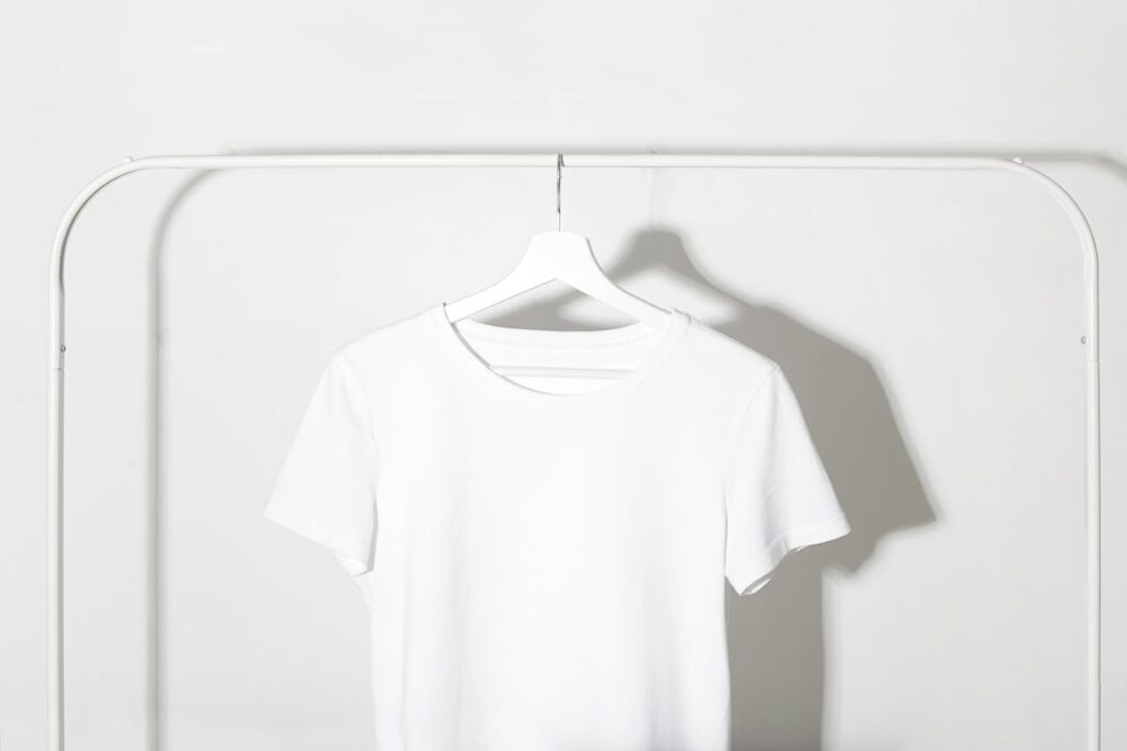 Plain white t-shirt on a hanger against a minimalist white backdrop with shadows.